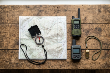 Two-way radio, compass on a topographic map, satellite messenger, and emergency whistle laid out on a wooden table for survival communication and navigation planning
